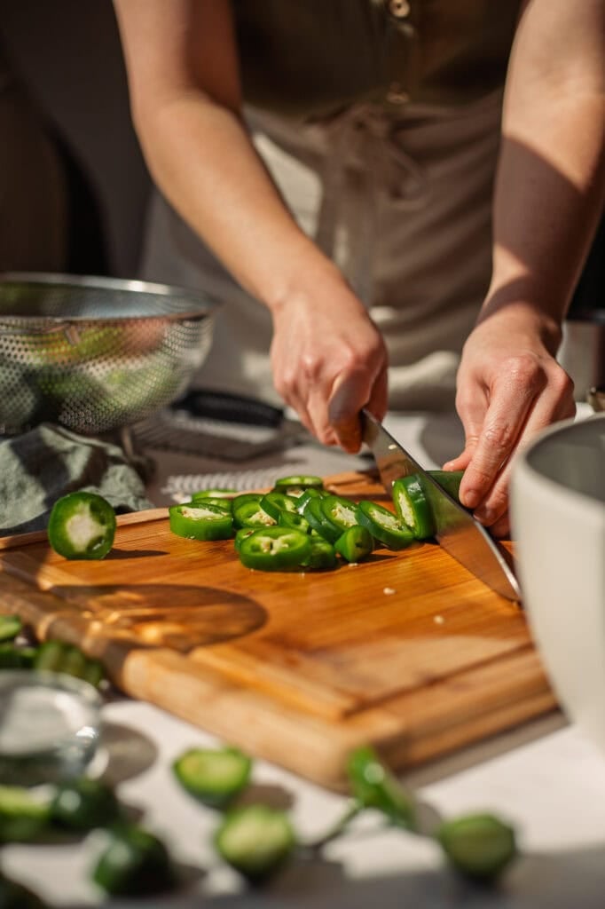 hands using a knife to cut jalapeños into slices on a wooden cutting board.